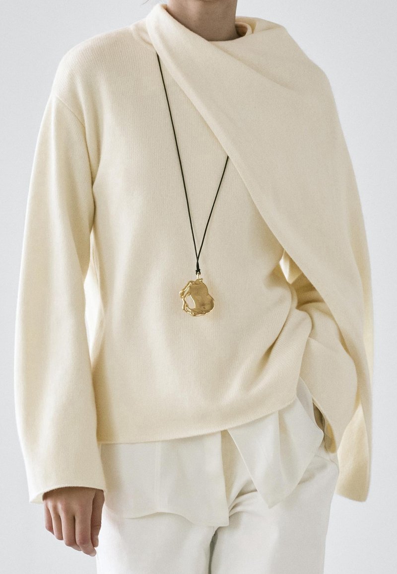 Personne portant un pull écru avec détail d’écharpe drapée, des couches blanches en dessous et un pendentif doré sur un collier en corde noire.