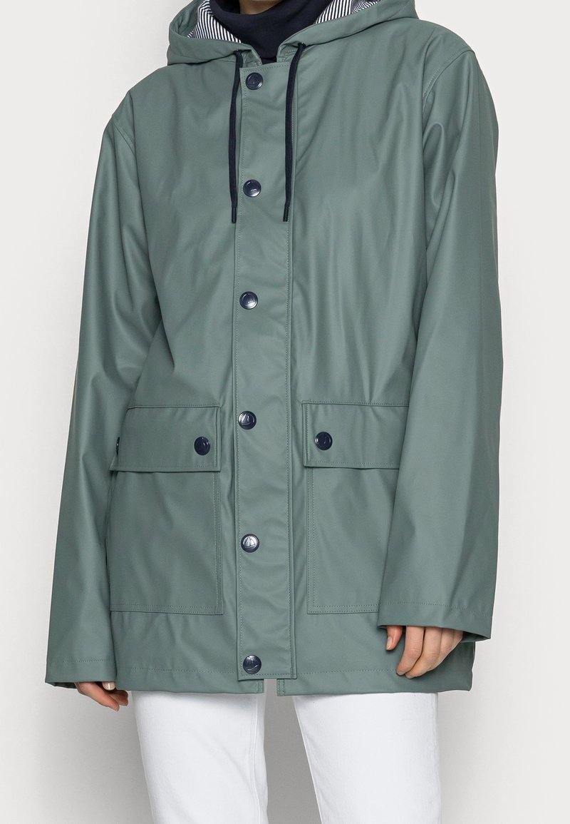 Vihreä hupparineuletakki, jossa on etunapit ja kaksi suurta läpällistä taskua, käytetty mustan paidan ja valkoisten housujen päällä.