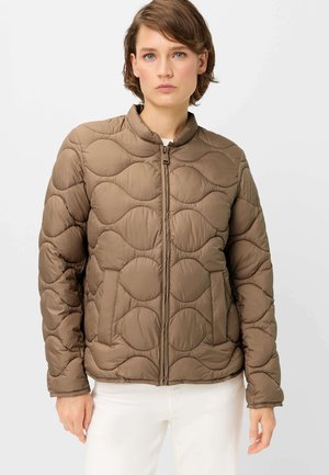 Woman with short brown hair wearing a brown quilted jacket with wave stitching and white pants, facing forward against a plain background.