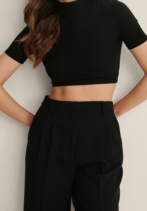 Vrouw draagt een zwart kort mouw crop top en hooggetailleerde zwarte broek, staand met handen in de heupen tegen een effen muur.