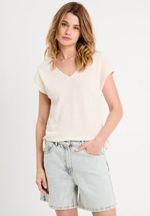 Młoda kobieta z włosami do ramion, ubrana w kremowy sweterek z krótkim rękawem i jasnoniebieskie dżinsowe szorty, ręka w kieszeni, na neutralnym tle.