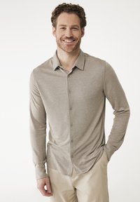 Chemise à manches longues en tissu beige clair, avec un col, un ourlet droit et une texture subtile. Associée à un pantalon de couleur claire.