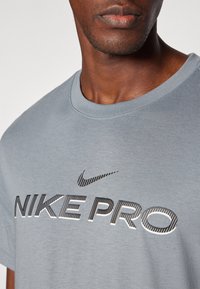 Grå Nike Pro T-skjorte med korte ermer, med en svart logo og tekstdesign på fronten. Myk tekstur, rund hals-type.