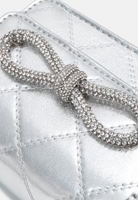Primo piano di una borsa in pelle metallizzata argento con dettaglio decorativo di un fiocco di strass sulla parte superiore.