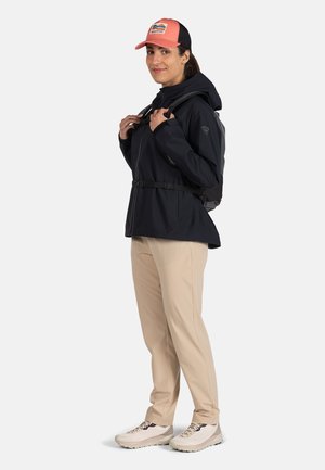 Giacca nera impermeabile con cappuccio regolabile e cintura, pantaloni beige, cappellino rosa e nero e scarpe da ginnastica chiare. Zaino sulla schiena.