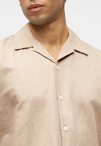 Lähikuva henkilöstä, jolla on päällään beige nappipaita leveällä kauluksella, näkyvissä kaula ja yläraaja.