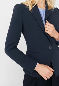 Blazer bleu marine en tissu structuré avec une fermeture à un bouton, un col à revers cranté et deux poches avant. Texture lisse avec une coupe ajustée.