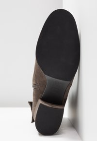 Semelle d'une chaussure habillée en daim marron avec talon en caoutchouc noir et semelle noire lisse, positionnée à la verticale contre un fond blanc d'angle.