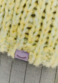 Pletená žltá čiapka s jemnou textúrou; má fialovú textilnú visačku s potlačeným dizajnom tváre. Pozadie je sivá vzorovaná tkanina.