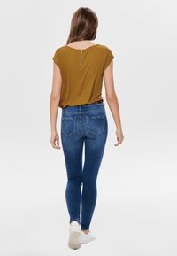 Senapsgul kortärmad topp med en metallblixtlåsdetalj på baksidan, tillsammans med kroppsnära mörkblå jeans och vita sneakers.