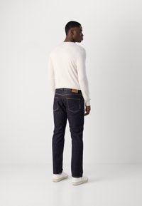 Tumed denim püksid kitsase lõikega, millel on kollane õmblus, tagumised taskud ja helepruun nahklabel. Mudel kannab valget pika varruka topi.