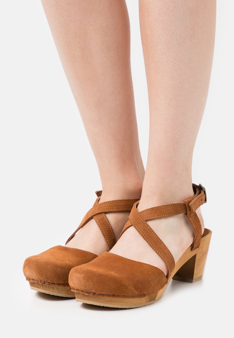 Sabots en daim brun à bout fermé avec talons en bois et brides croisées à la cheville, portés pieds nus sur un fond uni.