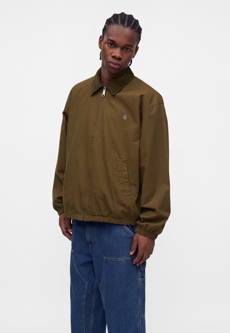 Jeune homme aux cheveux tressés, portant une veste zippée vert olive et un jean bleu ample, debout devant un fond blanc uni.