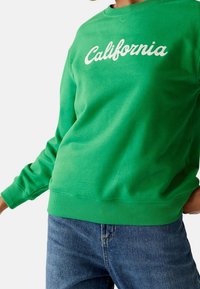 Felpa verde con scritta bianca "California". Presenta un collo rotondo e polsini a coste, realizzata in un tessuto morbido. Abbinata a jeans blu.