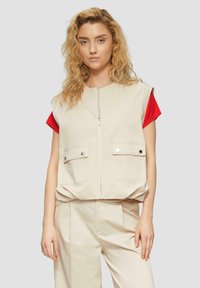 Beige ärmellose Weste mit zwei Fronttaschen, Reißverschluss und lockerer Passform, getragen über einem roten kurzärmeligen Hemd. Weicher Stoff, minimalistisches Design.