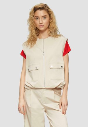 Beige mouwloze vest met twee voorzakken, ritsluiting en een losse pasvorm, gedragen over een rood shirt met korte mouwen. Zachte stof, minimalistisch ontwerp.