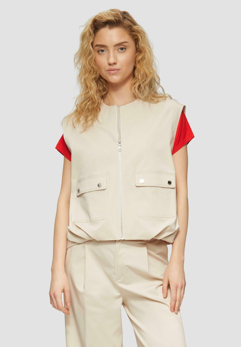 Beige ärmellose Weste mit zwei Fronttaschen, Reißverschluss und lockerer Passform, getragen über einem roten kurzärmeligen Hemd. Weicher Stoff, minimalistisches Design.