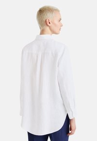 Camisa de lino blanca con mangas largas, cuello puntiagudo, pliegue en la espalda y dobladillo redondeado. Presenta puños abotonados y un corte holgado.