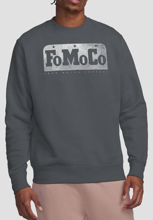 Mann trägt dunkelgraues Sweatshirt mit abgenutztem "FoMoCo Ford Motor Company"-Logo in weißem Text über der Brust.