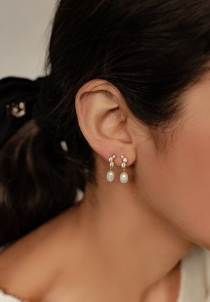 Goldene Tropfohrringe mit kleinen klaren Kristallen und weißen Perlen, die ein filigranes Design aufweisen, das kreisförmige Akzente über den Perlen beinhaltet.