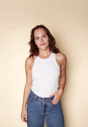 Weißes, geripptes Tanktop mit hohem Halsausschnitt; getragen mit hochtaillierten blauen Jeans aus Denim mit leichten Abnutzungen und kontrastierenden Nähten.