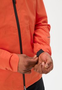 Persoon die de zwarte elastische manchetten van een feloranje jack met een rits aan de voorkant aanpast, tegen een effen witte achtergrond.