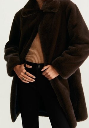 Cappotto in pelliccia sintetica marrone scuro indossato sopra un maglione beige a coste, abbinato a pantaloni neri a vita alta, con le mani che aggiustano la parte anteriore del cappotto.