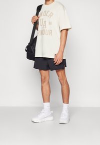 Krämfärgad T-shirt med kort ärm och beige text, svarta träningsshorts, vita sneakers och en svart väska. Enkelt design med en avslappnad passform.