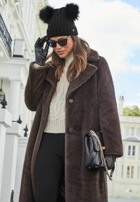 Abrigo de pelo sintético marrón, suéter de punto trenzado crema, gorro negro con dos pompones, guantes de cuero negros y bolso negro acolchado.