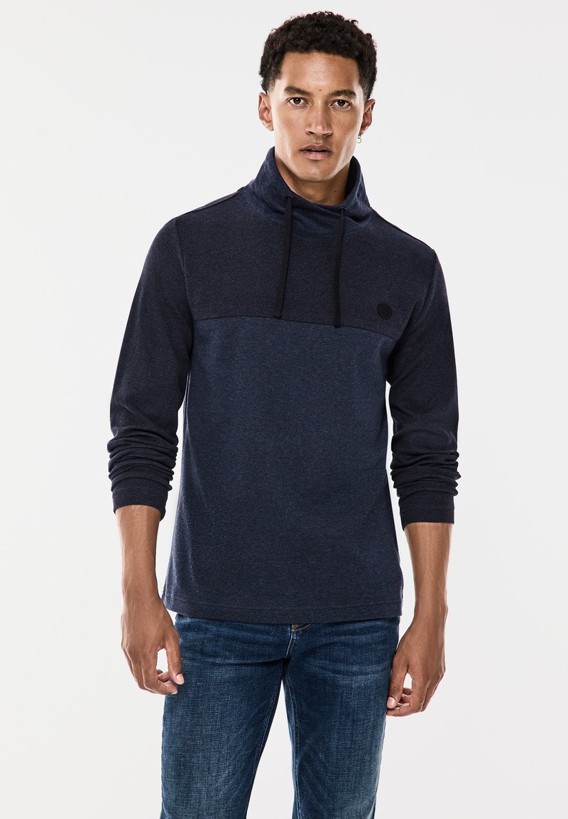 Námořnický modrý svetr s vysokým límcem, stahovací šňůrkou a kontrastní texturou na hrudi. Vyrobeno z měkké, texturované tkaniny.