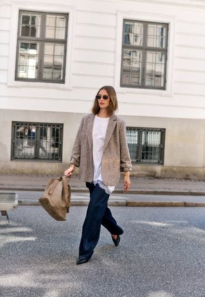 Femme portant des lunettes de soleil, un blazer beige, une chemise blanche et un pantalon foncé marchant dans la rue avec un grand sac beige devant un bâtiment blanc.
