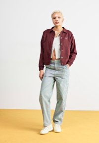 Casaco bordeaux, top crop listrado branco e calças listradas azul claro. As sapatilhas são de cor creme, e o fundo é liso, em amarelo claro.