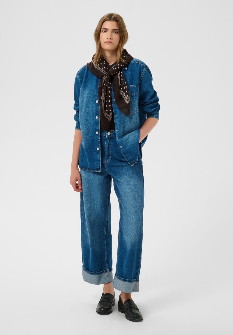 Femme portant une veste en jean bleue et un jean, une écharpe noire à pois, des mocassins noirs, une main dans la poche, debout devant un fond uni.