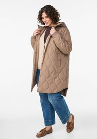 Pehmeäntakki beigen sävyssä, jossa on huppu, painonapit ja sivutaskut, päällä ruskea paita ja beige neuletakki, yhdistetty sinisiin farkkuihin ja ruskeisiin kenkiin.