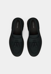 Sorte slip-on sko lavet af glat materiale, med flad sål, kvast-detaljer og kontrastsyninger langs kanterne.