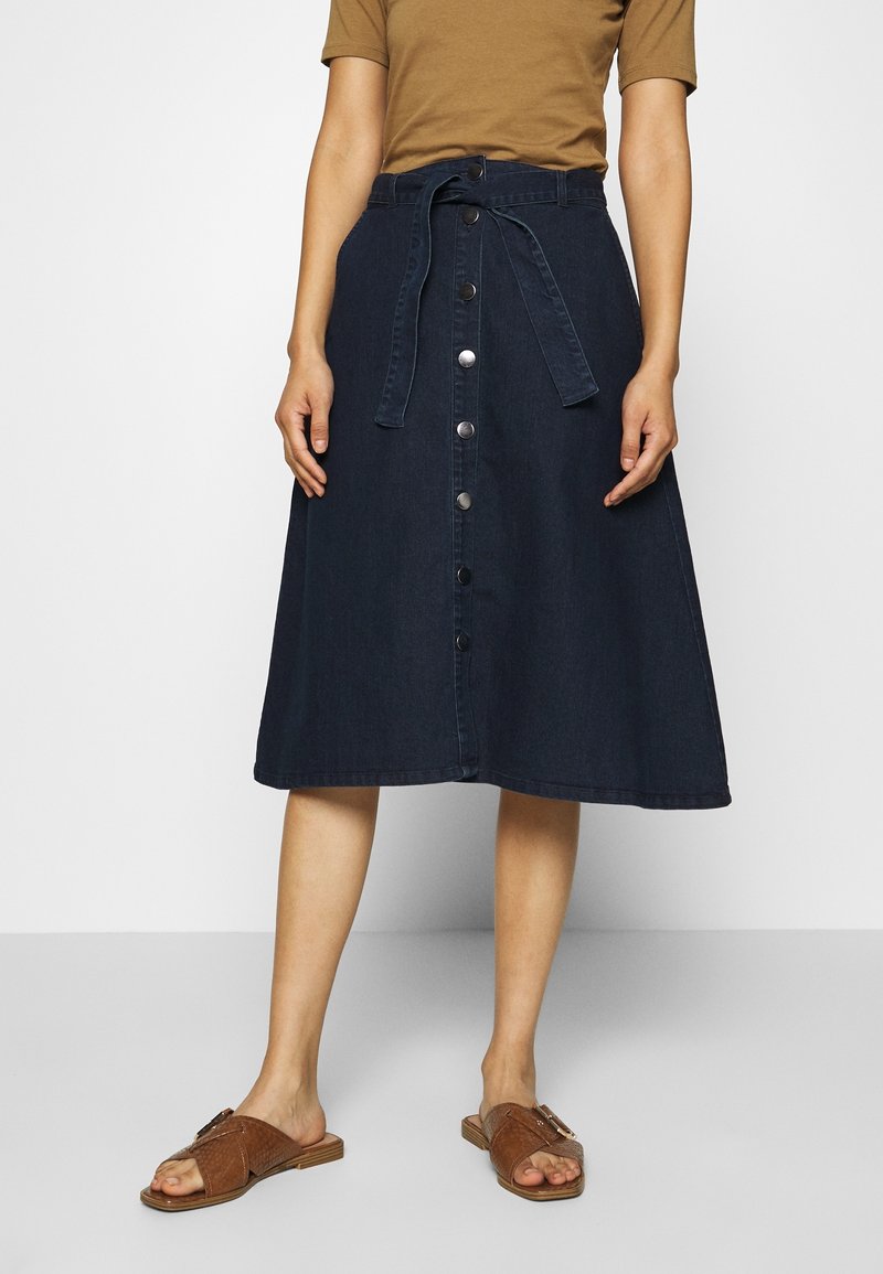 Jupe midi en denim bleu foncé avec une taille ceinturée, fermeture à boutons sur le devant et design évasé. Associée à des sandales à glisser bronzes.