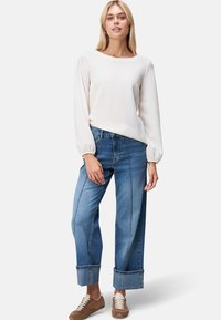 Weißer, strukturierter Pullover mit langen Ärmeln, kombiniert mit hellblauen, weit geschnittenen Jeans mit umgeschlagenen Säumen und sichtbaren Nähten. Braune Sneaker.