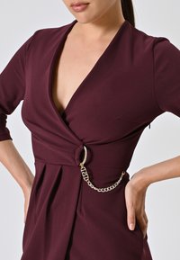 Bordeaux-rotes Kleid mit tiefem V-Ausschnitt und drei Viertelärmeln. Verfügt über ein gedrehtes Vorderteil und eine goldene Kettenakzentuierung an der Taille.