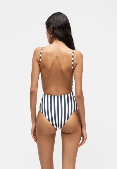 Femme aux longs cheveux foncés, vue de dos, portant un maillot de bain une pièce rayé vertical noir et blanc avec des bretelles fines et un dos ouvert.
