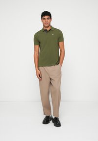 Olivově zelená polo triko s krátkým rukávem, jednobarevný design a kontrastní límec. Doplňuje béžové kalhoty a černé boty.