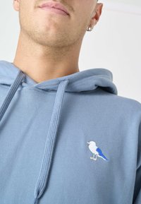 Felpa leggera blu chiaro in morbido tessuto con un design di uccello ricamato in bianco e blu sul petto. I laccetti completano il look.