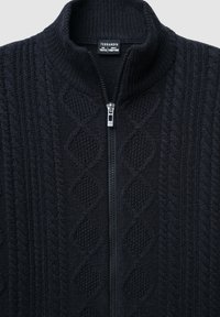 Ciemnoniebieski dzianinowy sweter z zamkiem błyskawicznym, z wysokim kołnierzem, teksturowanym wzorem w warkocze i srebrnym zamkiem. Etykieta marki widoczna wewnątrz.