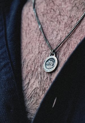 Silver pendant on a twisted chain, featuring an intricate engraved design, resting on a hairy chest with a dark fabric background.
