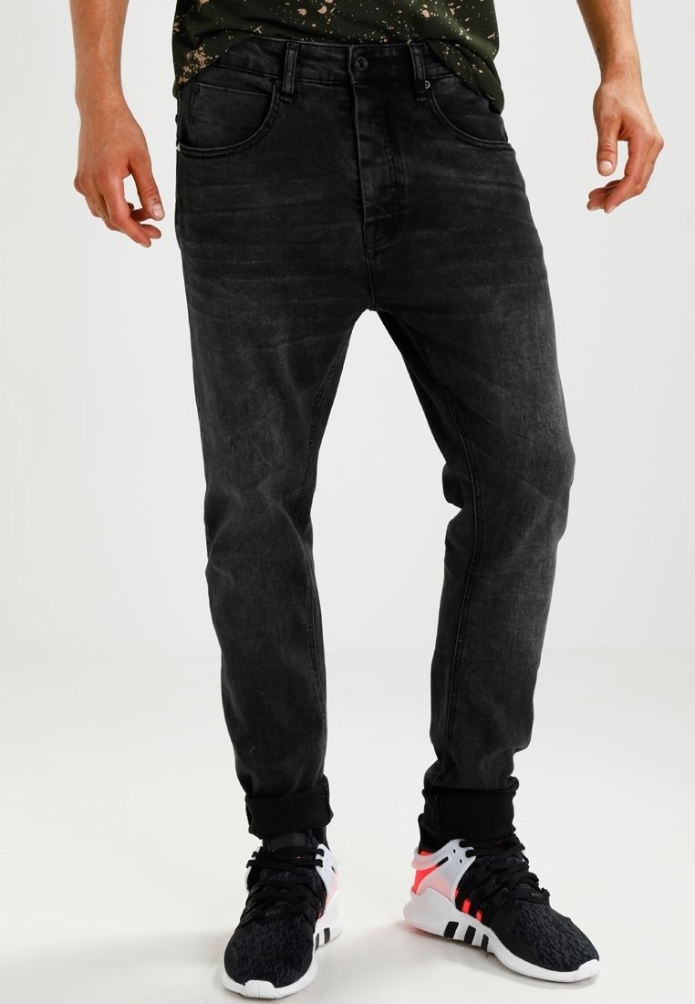 Hombre con vaqueros negros desgastados doblados en los puños y zapatillas negras y blancas con detalles en rojo, de pie frente a un fondo liso.