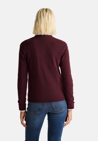 Burgunderfarbener Strickpullover mit rundem Halsausschnitt und gerippten Bündchen, getragen über blauen Jeans. Der Pullover hat eine glatte Textur und eine taillierte Passform.