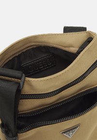 Sac en toile beige en bandoulière avec des zips noirs, une sangle réglable et un logo GUESS visible à l'intérieur ainsi que sur la poche avant.