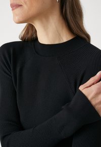 Svart ribbestrikket genser med høy hals og lange ermer med teksturdetaljer. Nærbilde viser glatt stoff og avslappet passform.