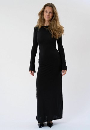 Femme aux cheveux longs et ondulés portant une robe noire longue ajustée et des chaussures noires pointues, debout devant un fond clair uni.