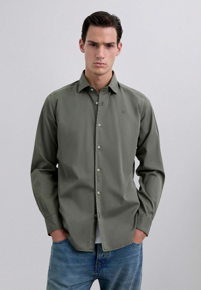 Camisa de botón verde oliva hecha de tela suave, con un cuello clásico y un sutil bordado de logo, combinada con unos vaqueros de mezclilla de color azul claro.