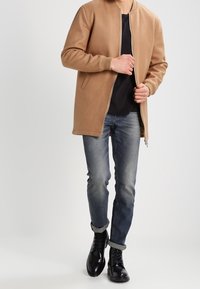 Tans zip-up jacka med ribbade muddar, stylad över en svart skjorta, i kombination med bleka blåjeans och svarta snörboots. Slät textur.